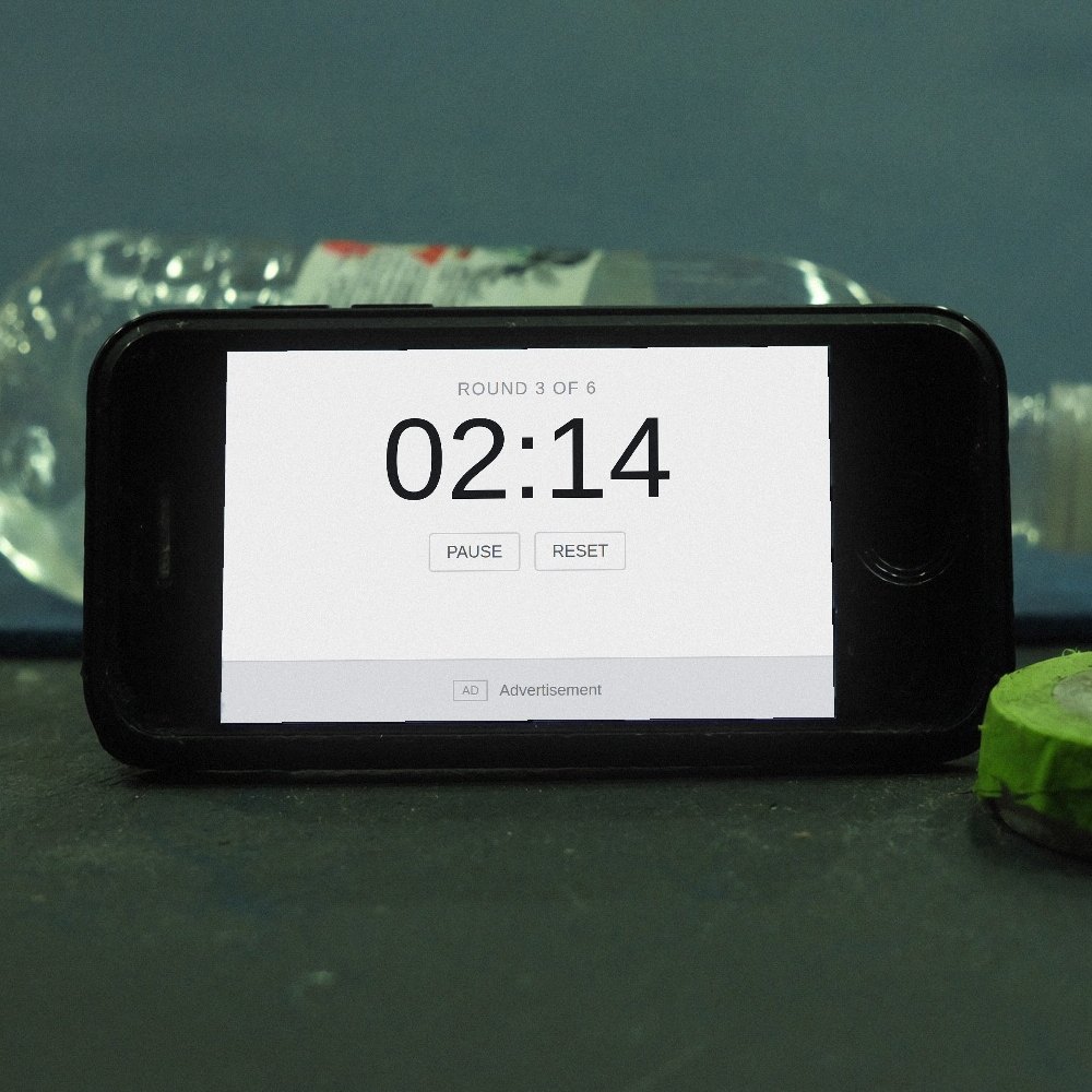 A scuffed phone propped against a water bottle at the edge of a gym mat, running a plain white countdown app with an advert along the bottom of it